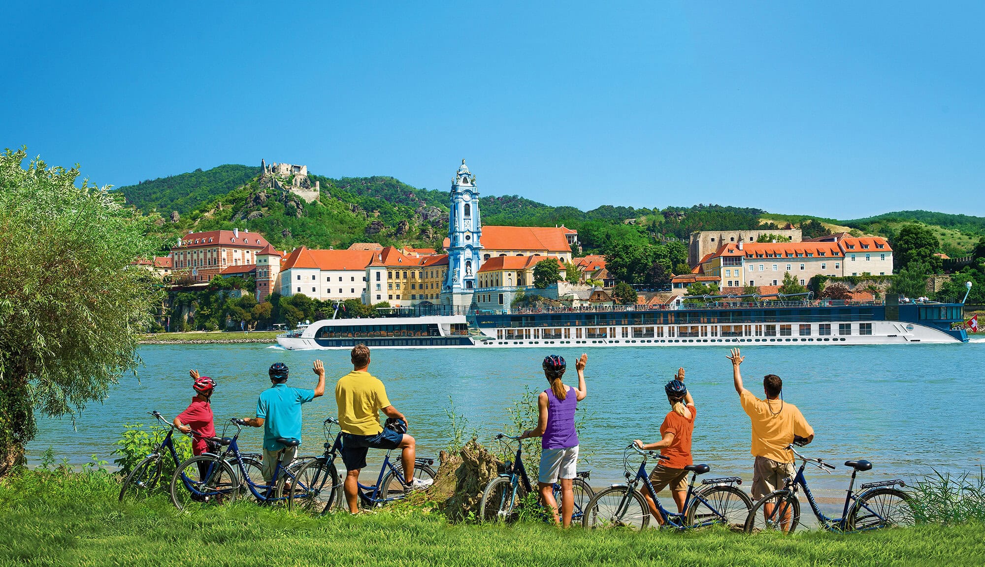 Three-generation family on the riverbank waving at a luxury river cruise—capturing the spirit of river cruises for families across Europe.
