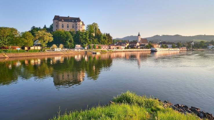 A peaceful Rhine River scene with a hilltop castle above a small riverside town and church. Calm water reflects the buildings trees and warm evening light along one of the best European rivers to cruise.
