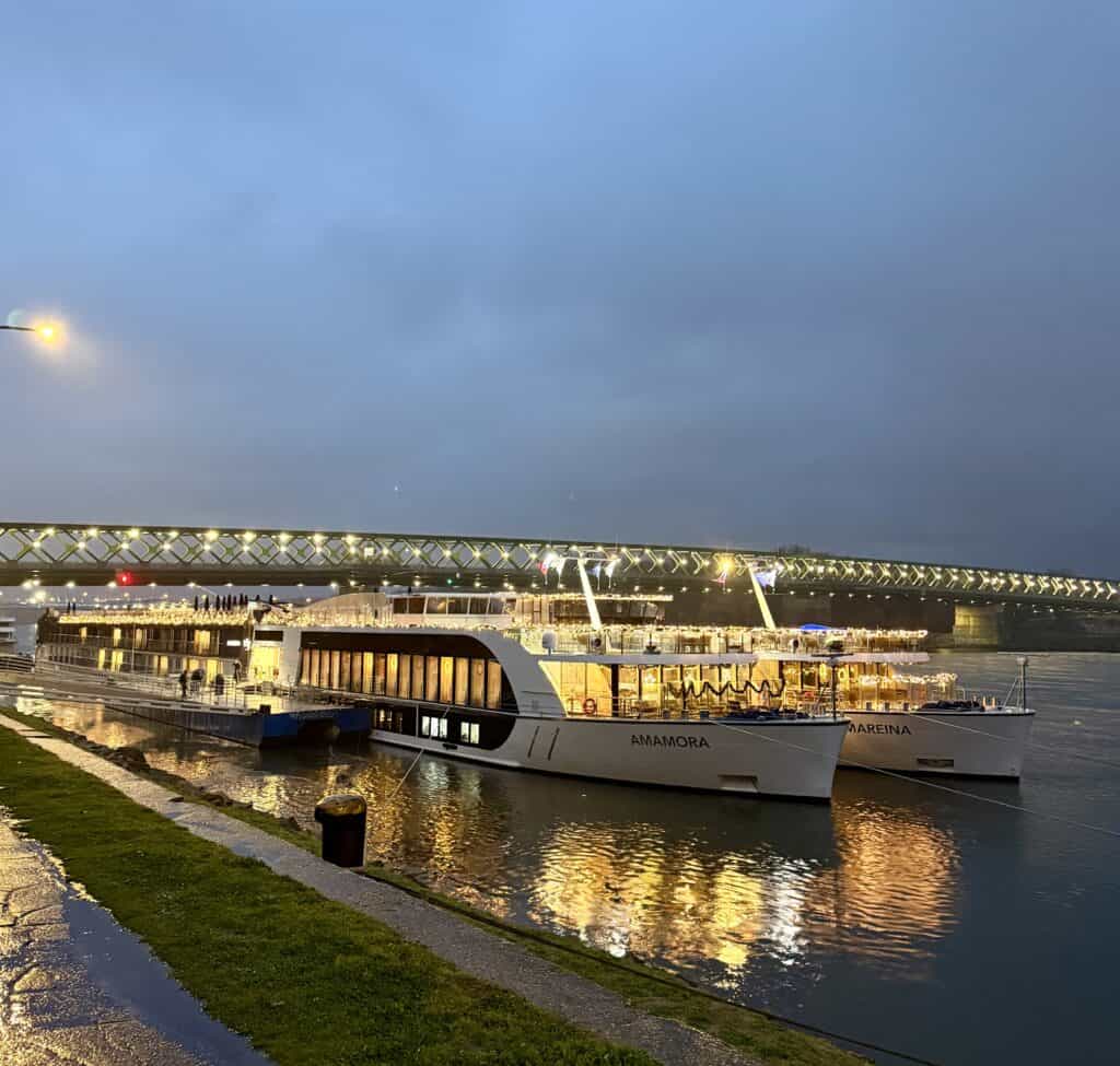 AmaMora and AmaReina river cruise ships dock side by side at dusk with warm lights glowing across their decks. The two AmaWaterways ships offer a visual reference for travelers comparing Tauck vs AmaWaterways river cruises.