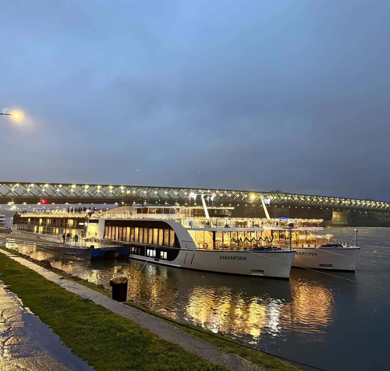 AmaMora and AmaReina river cruise ships dock side by side at dusk with warm lights glowing across their decks. The two AmaWaterways ships offer a visual reference for travelers comparing Tauck vs AmaWaterways river cruises.