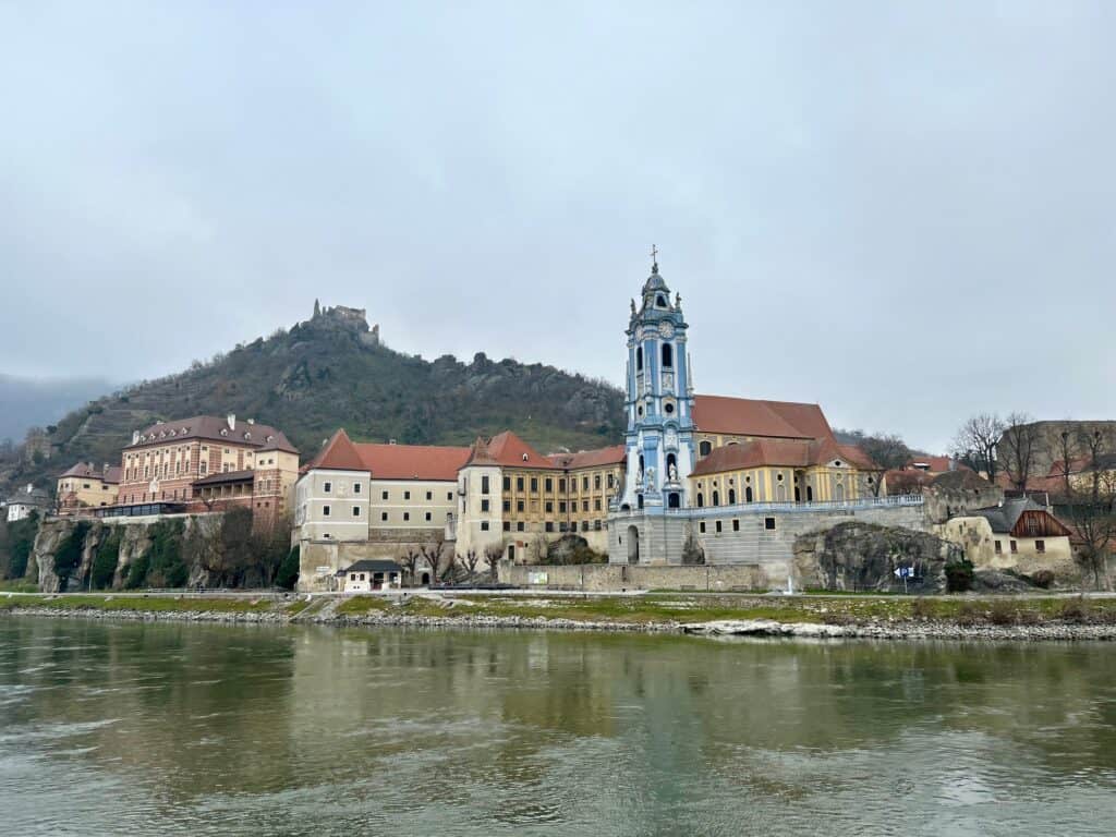 The blue tower of Dürnstein Abbey rises above the Danube River with the ruins of Dürnstein Castle on the hillside behind it under a cloudy sky. This scenic stop highlights the historic villages you can experience on a Danube vs Rhine River Cruise.