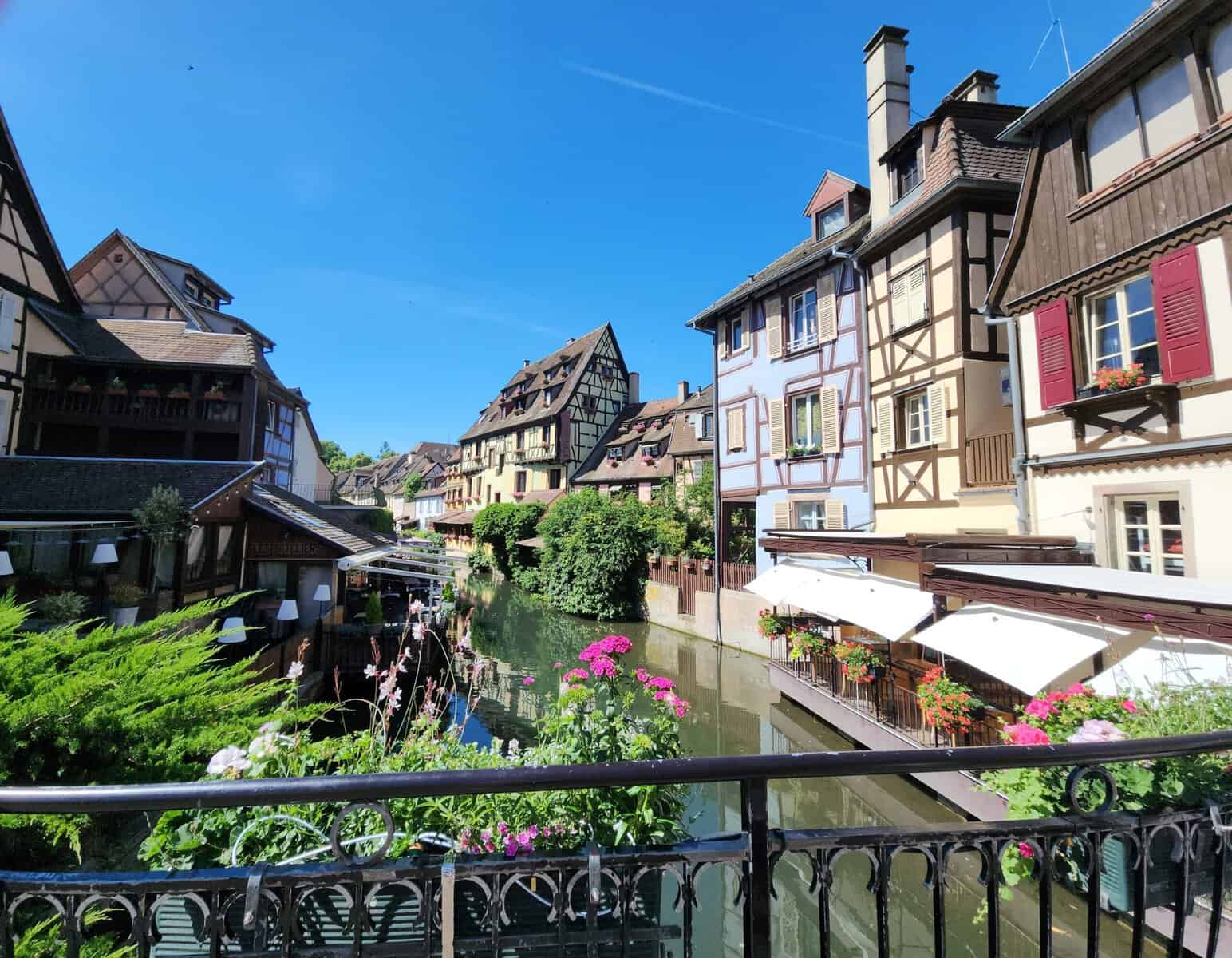 Colorful half timbered houses with wooden shutters and flower boxes line a narrow canal in Colmar France. Waterfront restaurants with white awnings overlook the water beneath a bright blue sky.