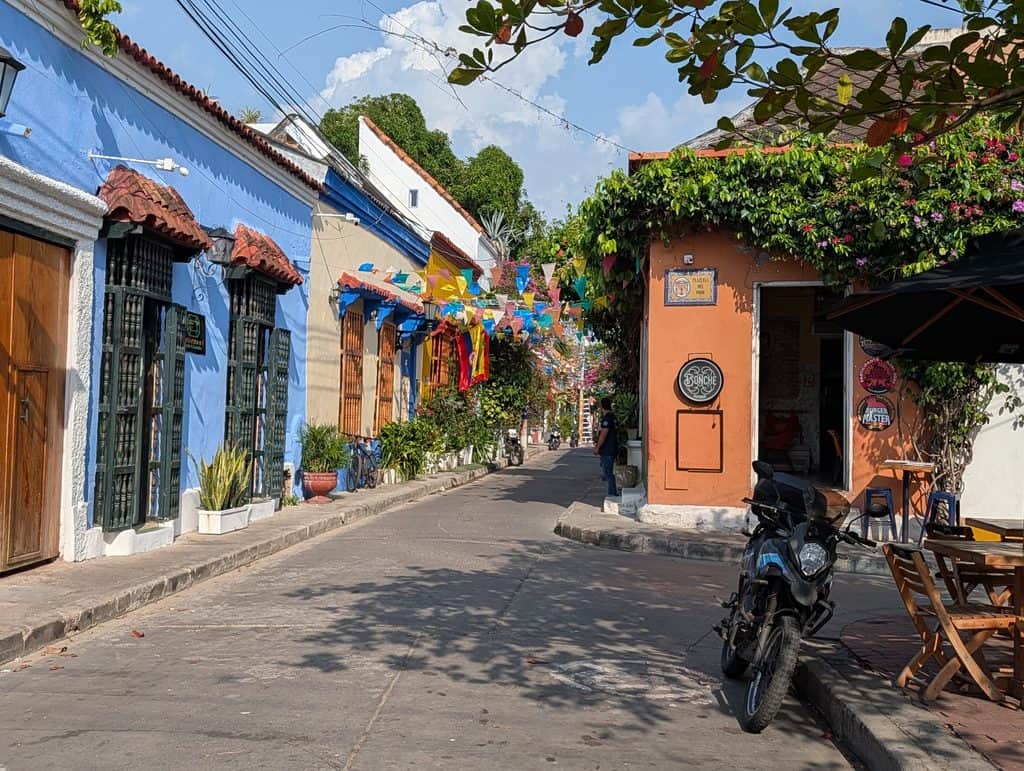A sunny narrow street lined with colorful blue orange and white buildings decorated with plants and strings of bright flags. A motorcycle is parked beside outdoor tables along the quiet street.