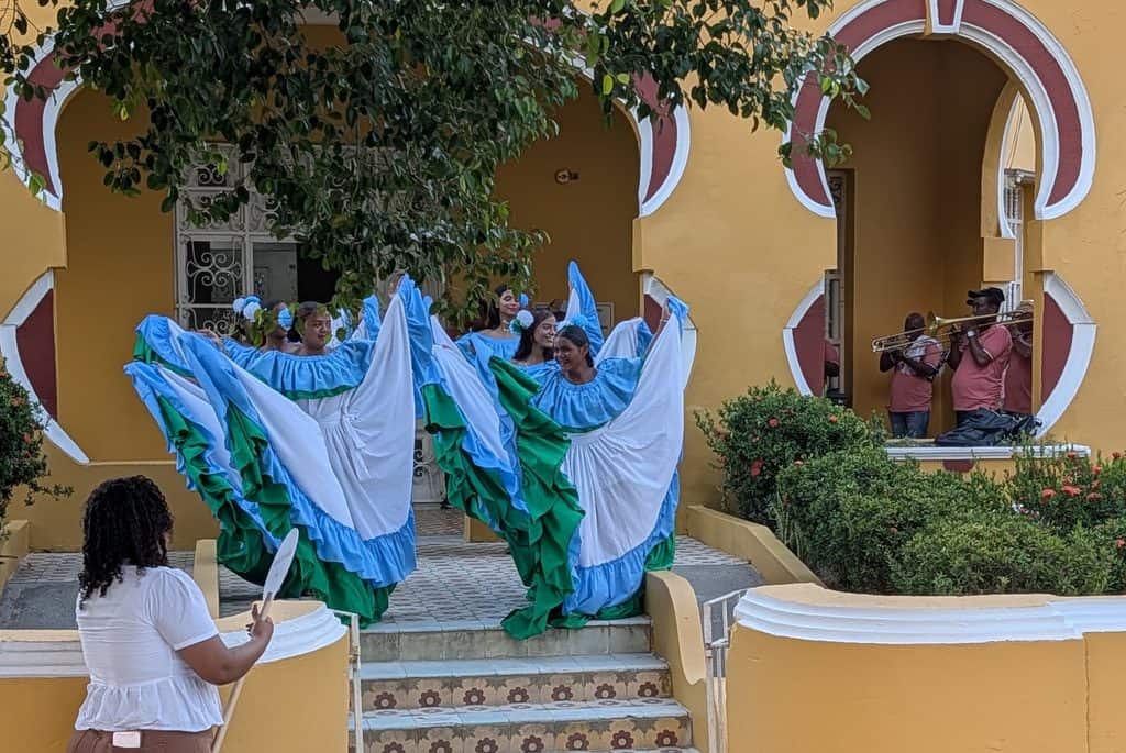 Dancers in flowing blue green and white dresses perform beside musicians playing brass instruments in front of a yellow building with large arched doorways. The colorful performance highlights local culture experienced during a Colombia River Cruise.