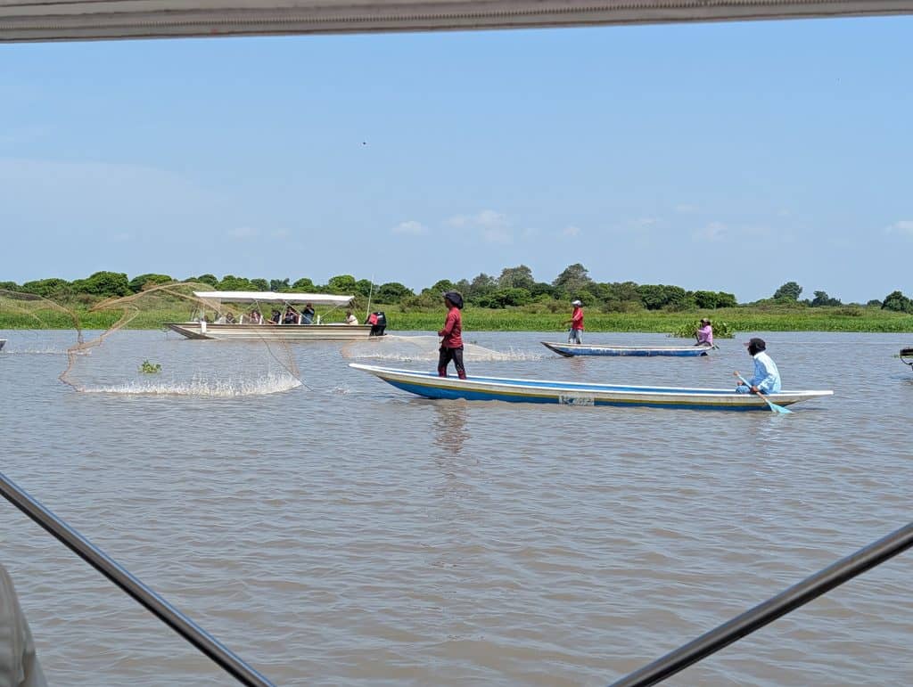 Fishermen stand in long narrow wooden boats while throwing circular fishing nets across the river. A covered passenger boat passes behind them near the lush green shoreline during a Colombia River Cruise.