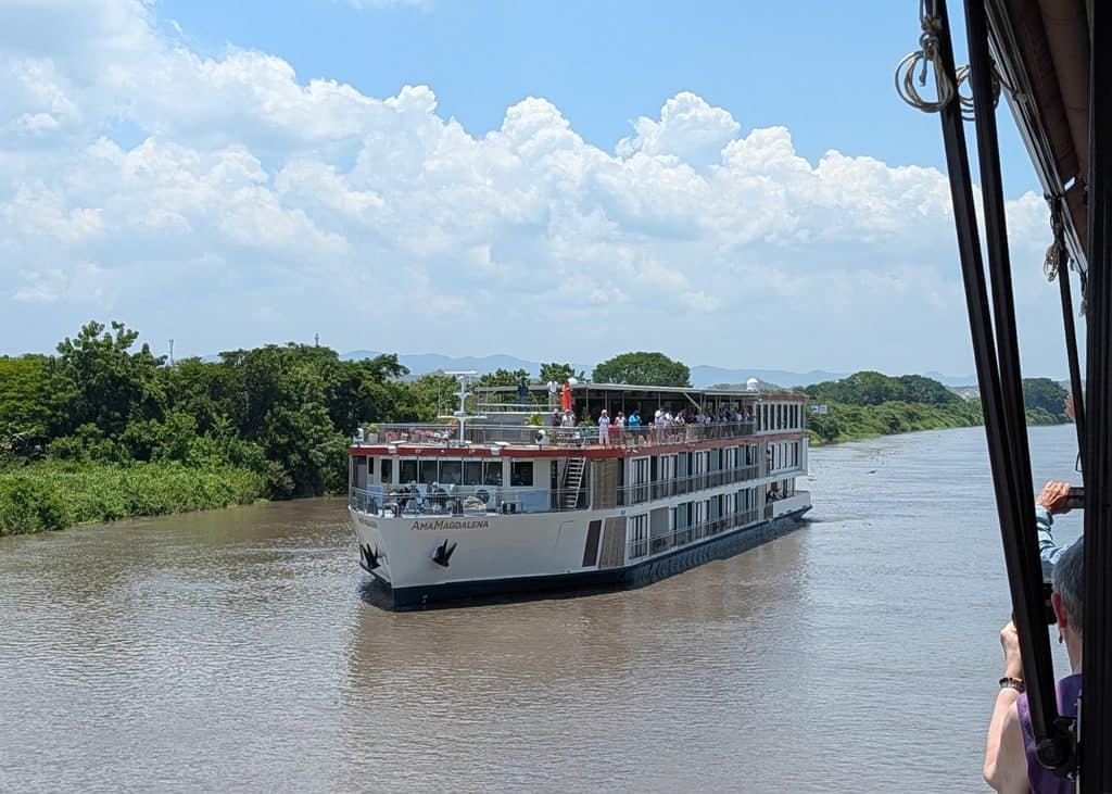The AmaMagdalena river cruise ship sails along a wide brown river bordered by dense green vegetation under a bright blue sky. Passengers gather on the upper deck as the ship approaches.