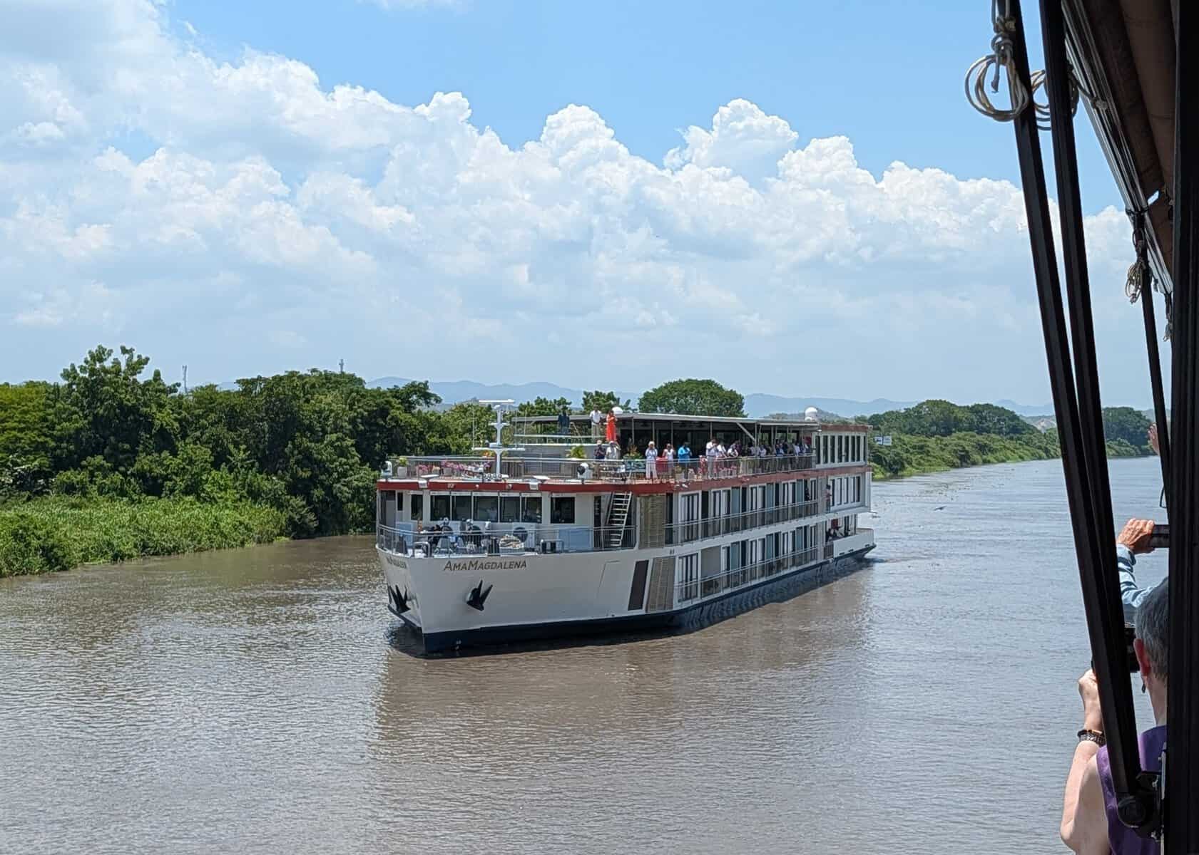 The AmaMagdalena river cruise ship sails along a wide brown river bordered by dense green vegetation under a bright blue sky. Passengers gather on the upper deck as the ship approaches.
