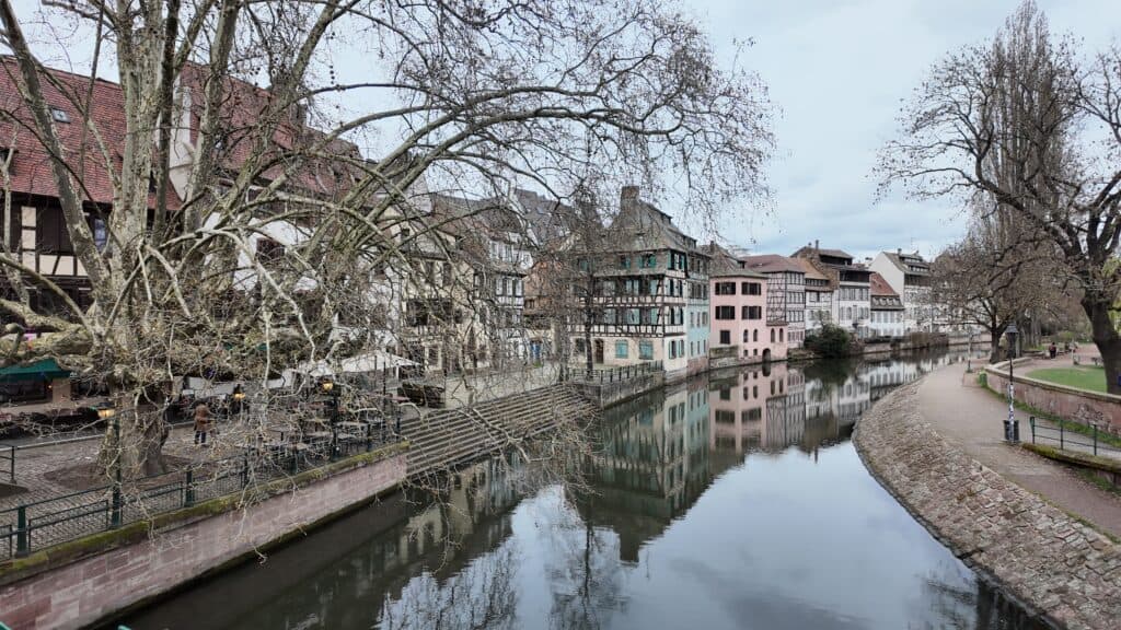 Quiet canal running between historic half timbered European buildings and a tree lined walking path on an overcast day. The traditional architecture reflects the historic towns travelers can explore during a European river cruise.
