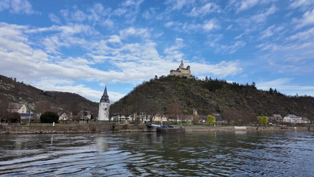 Historic hilltop castle overlooking the Rhine River above a small village with a church steeple, viewed from a European river cruise on a bright day with blue skies.