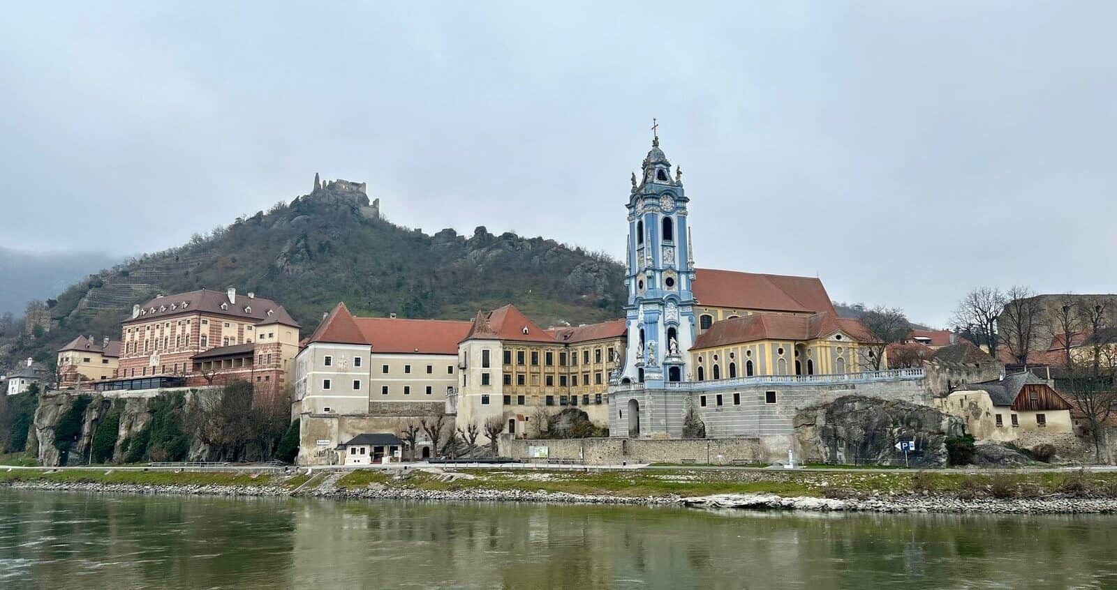 A scenic view along a European river cruise, showcasing Dürnstein, Austria, with the iconic blue Baroque tower of the Dürnstein Abbey and hillside vineyards overlooking the Danube River.