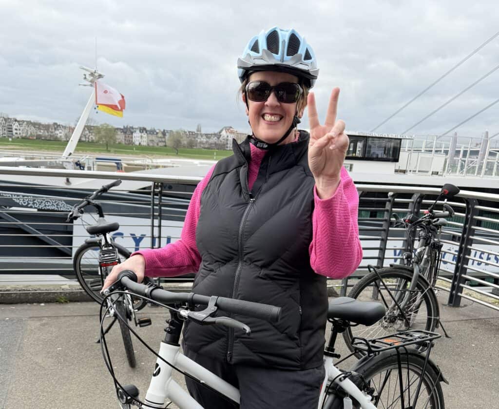 A woman wearing a bicycle helmet stands beside a rental bike along the Rhine River in Düsseldorf while giving a peace sign. Active excursions like cycling through riverside cities are common on a Rhine River cruise.