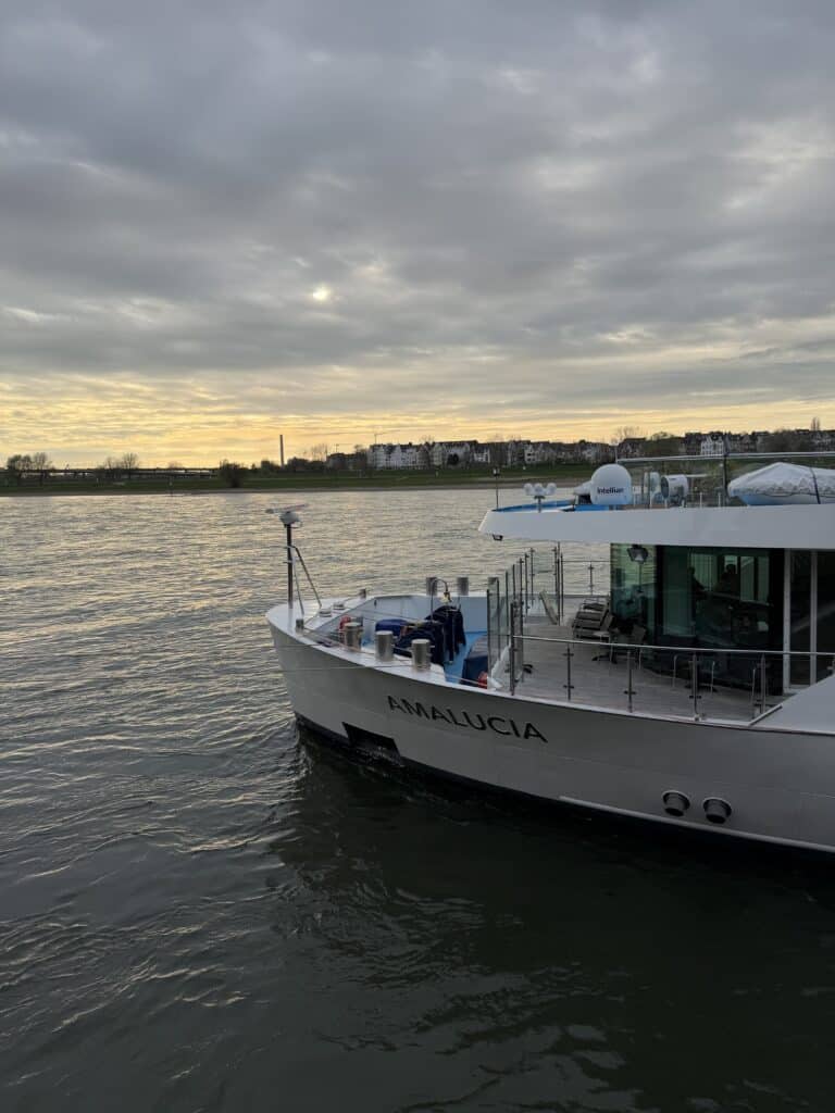 White river cruise ship docked along a calm river beneath a dramatic sunset sky with layered clouds and soft golden light reflecting on the water.