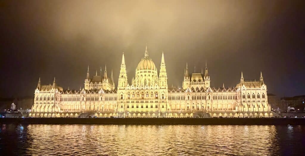 The Hungarian Parliament Building glows with golden lights along the Danube River in Budapest at night. Its grand central dome and Gothic Revival towers reflect across the dark water beneath a cloudy sky.