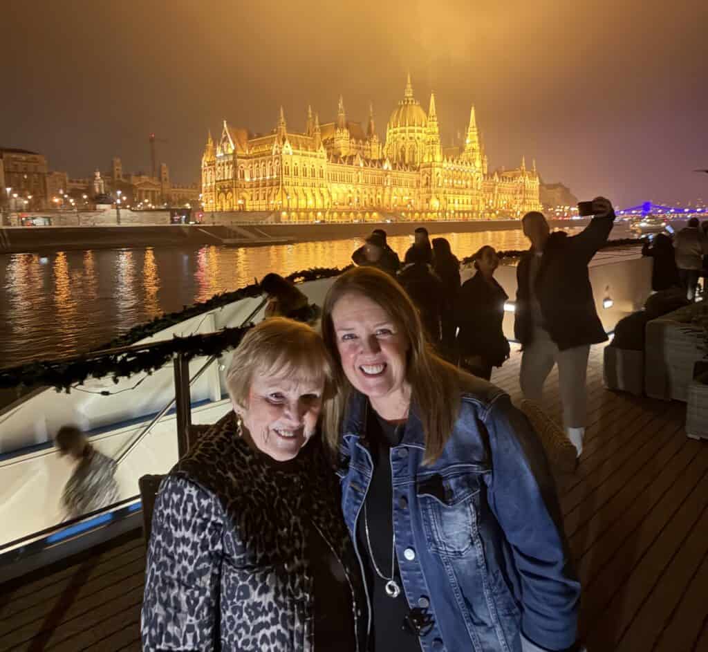 Two women stand on the deck of a river cruise ship with the illuminated Hungarian Parliament Building reflecting across the Danube River in Budapest at night. This iconic view is one of the signature experiences when comparing a Danube vs Rhine River Cruise.