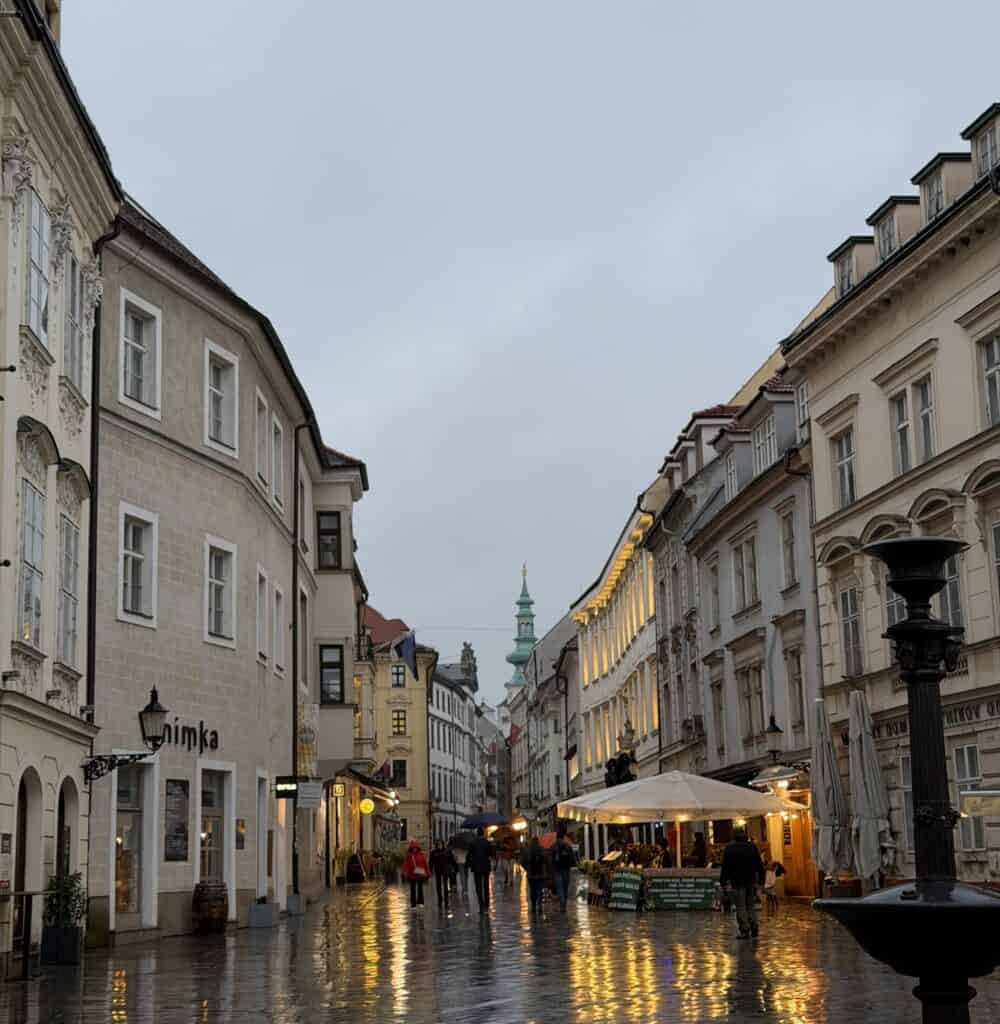 A rain-soaked street in Bratislava's Old Town is lined with historic buildings, cafés, and glowing lights reflected on the cobblestones as people walk with umbrellas. Bratislava is one of the charming cities visited on many Danube River cruises.
