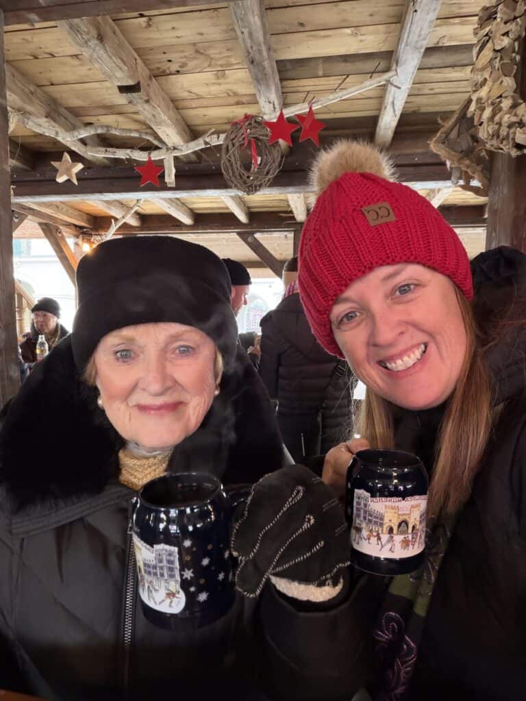 Two women smile while holding steaming mugs at a festive Christmas market in Munich decorated with rustic wooden beams and red star ornaments. Christmas market excursions are a popular extension to both Danube vs Rhine River Cruise itineraries during the holiday season.