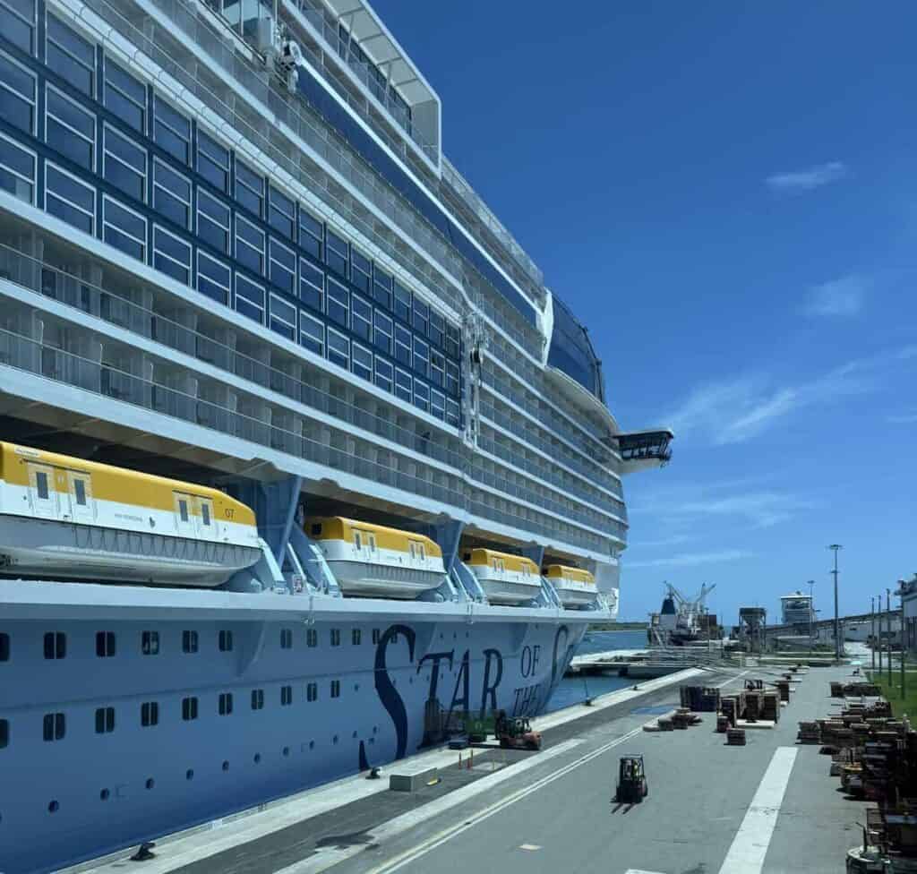 Star of the Seas docked at a cruise terminal under a bright blue sky with rows of balcony cabins and yellow lifeboats visible along the side of the massive ship.