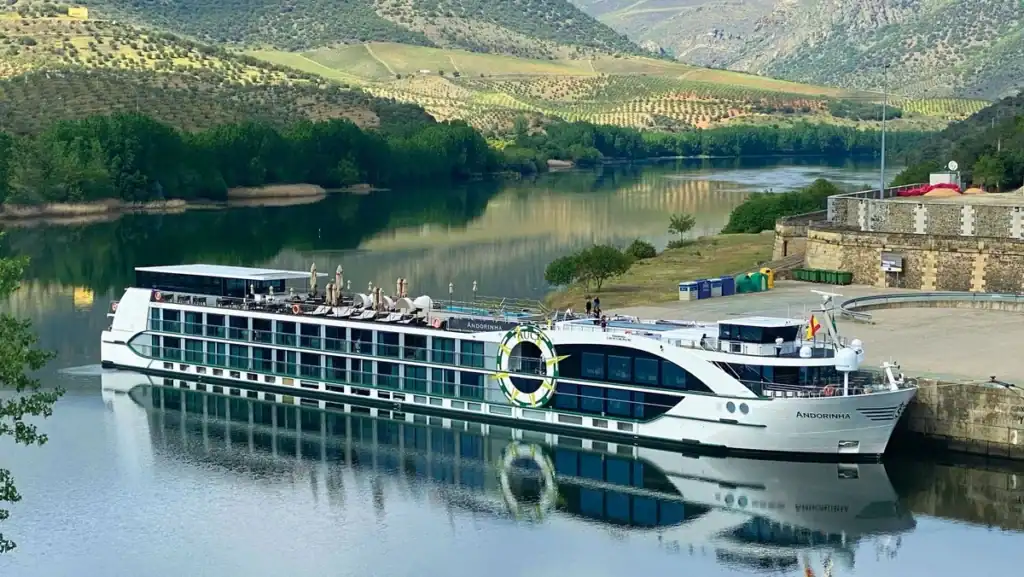 Tauck Andorinha river cruise ship docked on calm water surrounded by green hills and terraced farmland. The ship and peaceful landscape show the river cruising experience relevant to a Tauck vs. Viking comparison.