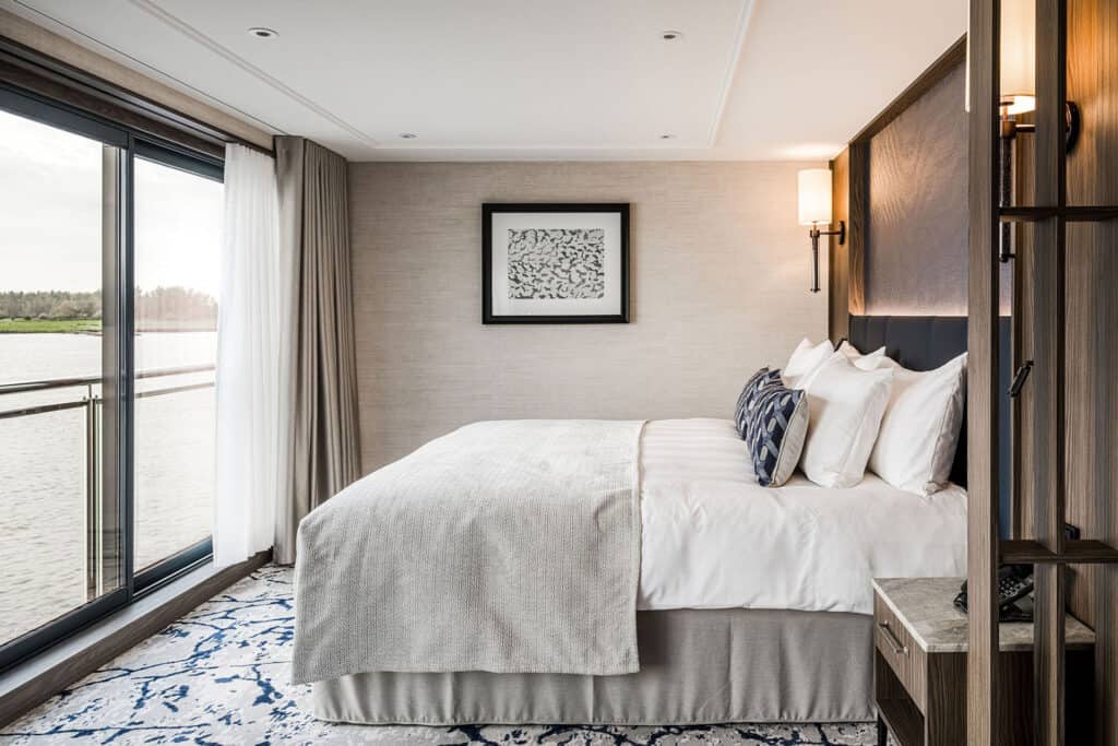 Modern river cruise cabin with a large bed beside floor to ceiling sliding glass doors overlooking the river. The bright room has neutral decor with white bedding and blue accents.