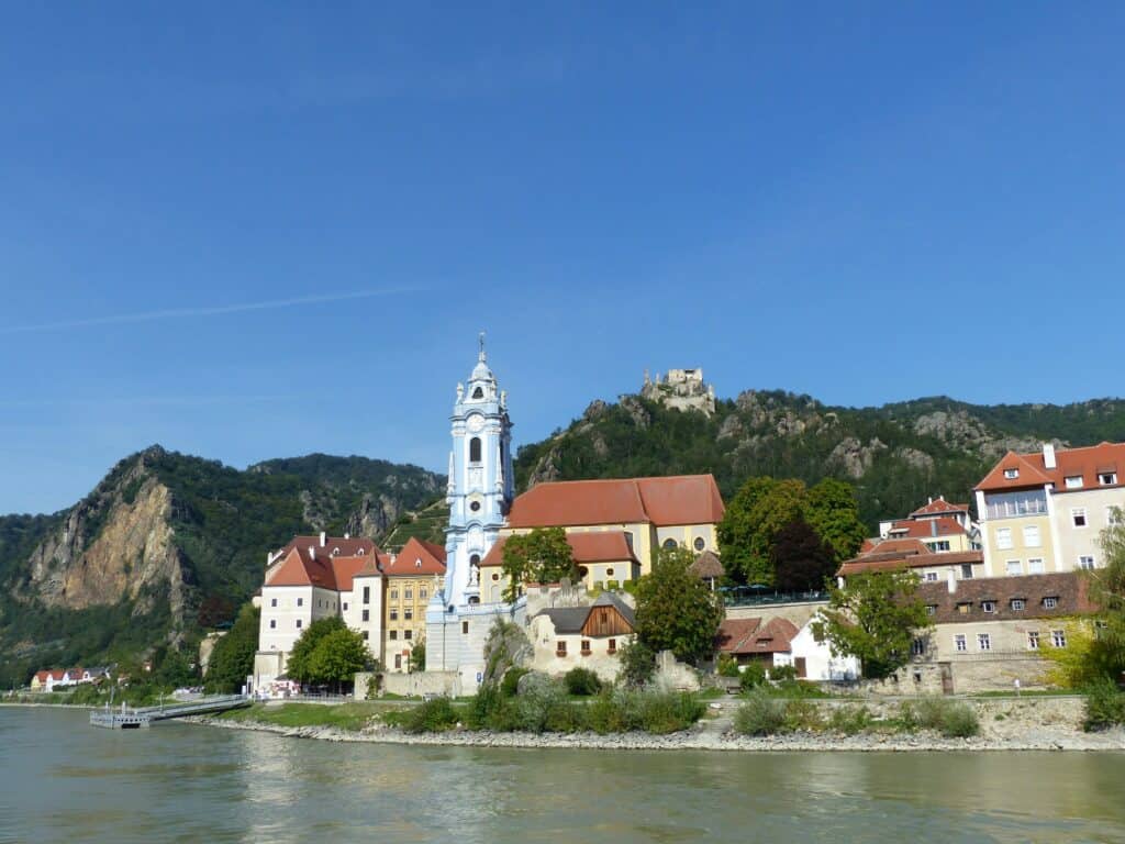 A historic European riverside village features a striking blue church tower, colorful rooftops, and castle ruins overlooking the river beneath a clear blue sky. This scenic destination is a memorable stop on a European river cruise, offering a blend of history, culture, and beautiful landscapes.