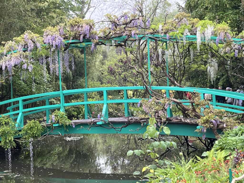 A turquoise arched footbridge crosses a quiet pond beneath cascading purple and white wisteria. Dense green foliage surrounds the bridge and reflects in the dark water.