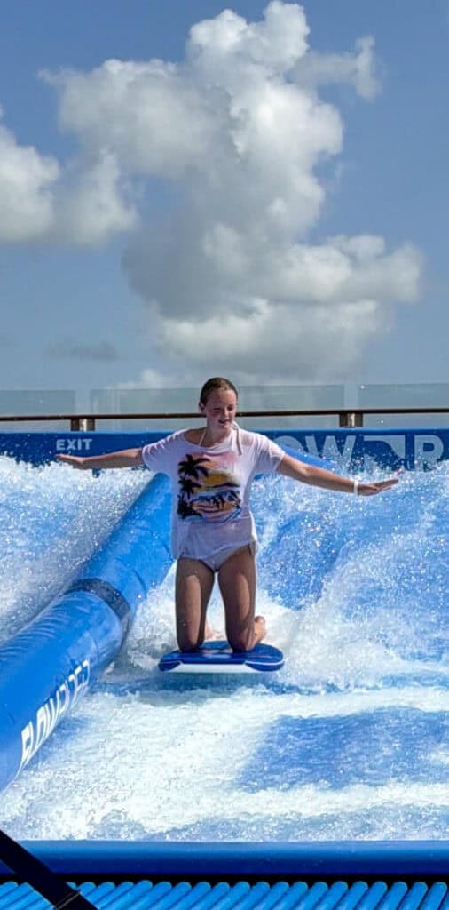 A teenager kneels on a blue board while balancing on rushing water in a cruise ship surf simulator beneath a bright blue sky. The attraction displays the words "FLOWRIDER" and "EXIT" beside the artificial wave.