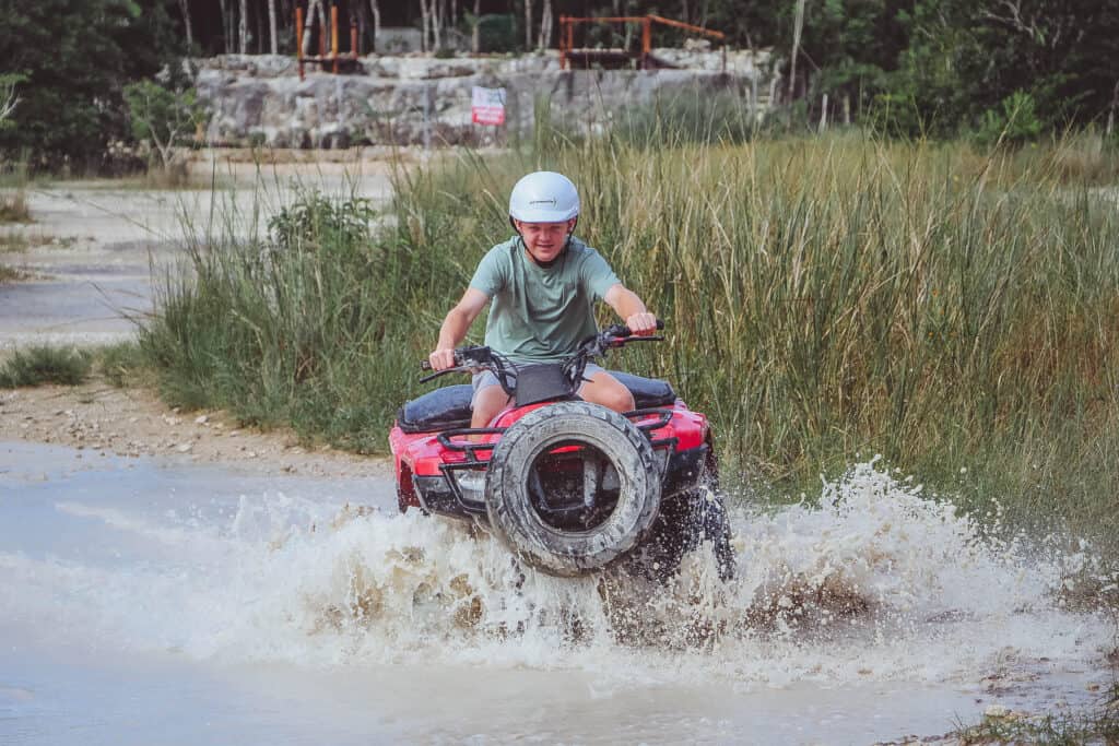 A helmeted teenager rides a red ATV through a large muddy puddle while water splashes around the vehicle. Tall grasses and trees surround the off road trail during a cruise excursion.