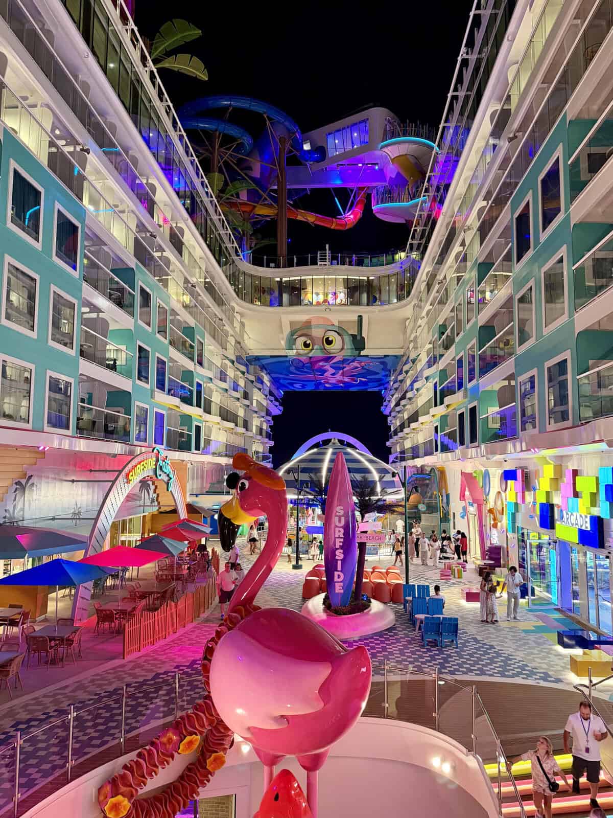 A colorful open air neighborhood on a mega cruise ship at night with twisting waterslides overhead and a large pink flamingo sculpture in the center. Bright signs read "SURFSIDE" and "ARCADE" as passengers walk between restaurants seating areas and family attractions.