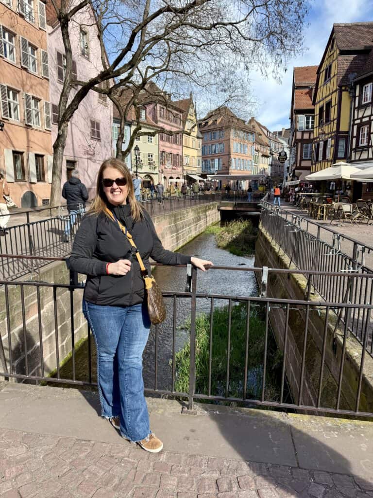 A woman traveling alone smiles on a small bridge over a canal surrounded by colorful historic buildings and outdoor cafes. The sunny European streetscape shows the freedom to explore destinations independently during a river cruise.