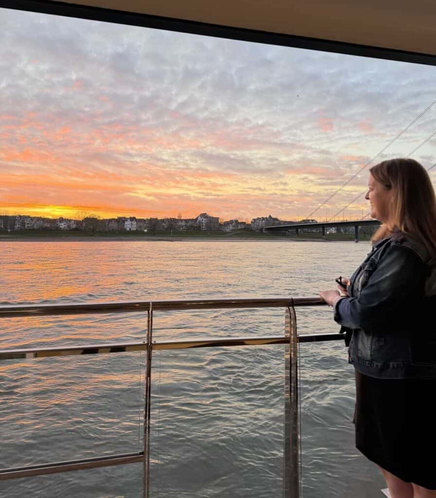 A woman stands on the outdoor deck of a river cruise ship watching a vivid orange and pink sunset reflect across the water. The peaceful evening scenery captures the quiet independence of traveling on a solo river cruise.