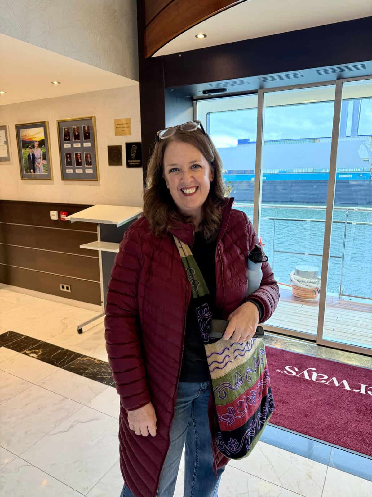 A solo traveler smiles inside the entrance of a river cruise ship dressed for a day of exploring with a coat scarf bag and water bottle. Glass doors behind her overlook the river and dock.