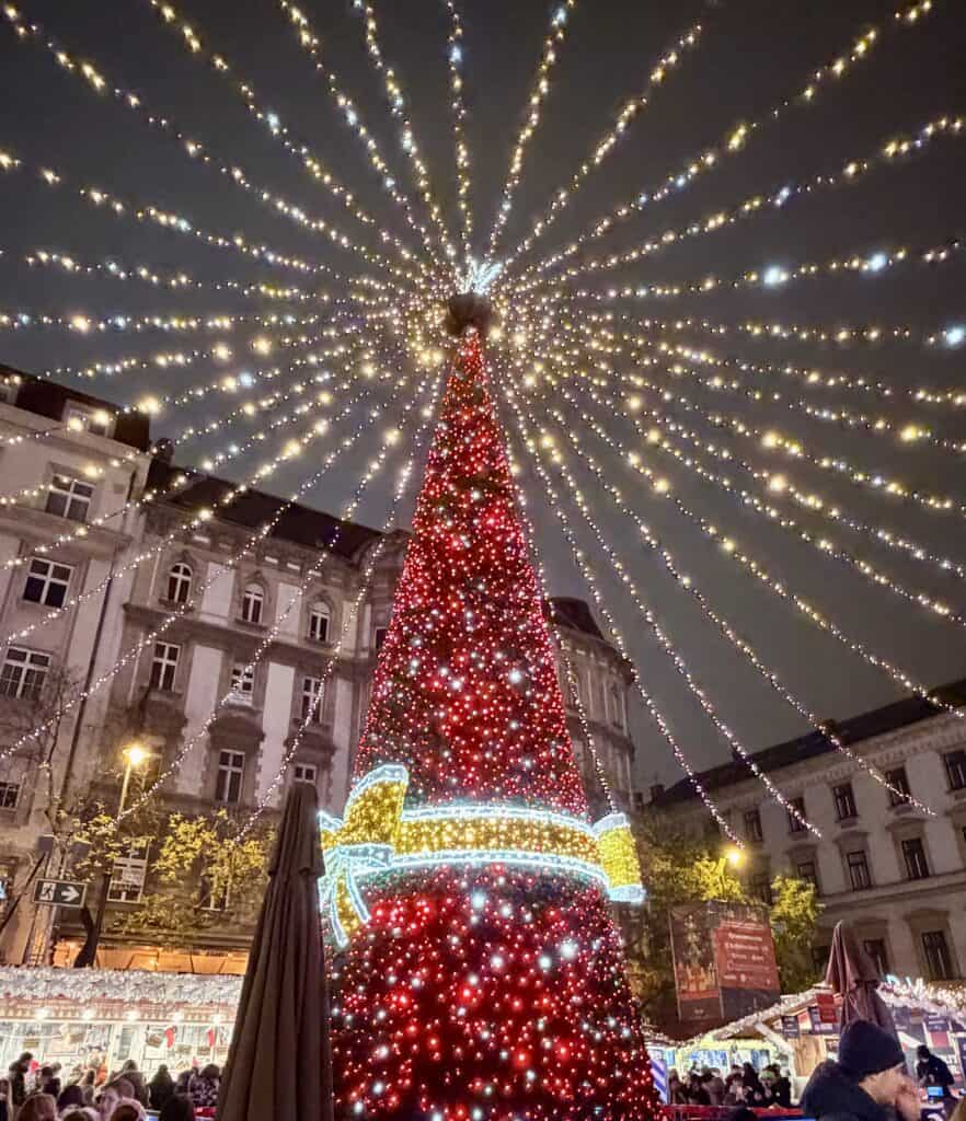 A towering Christmas tree covered in red and white lights stands beneath a canopy of twinkling string lights at a European Christmas market. A glowing ribbon decoration wraps around the tree while festive market stalls and visitors fill the square below.
