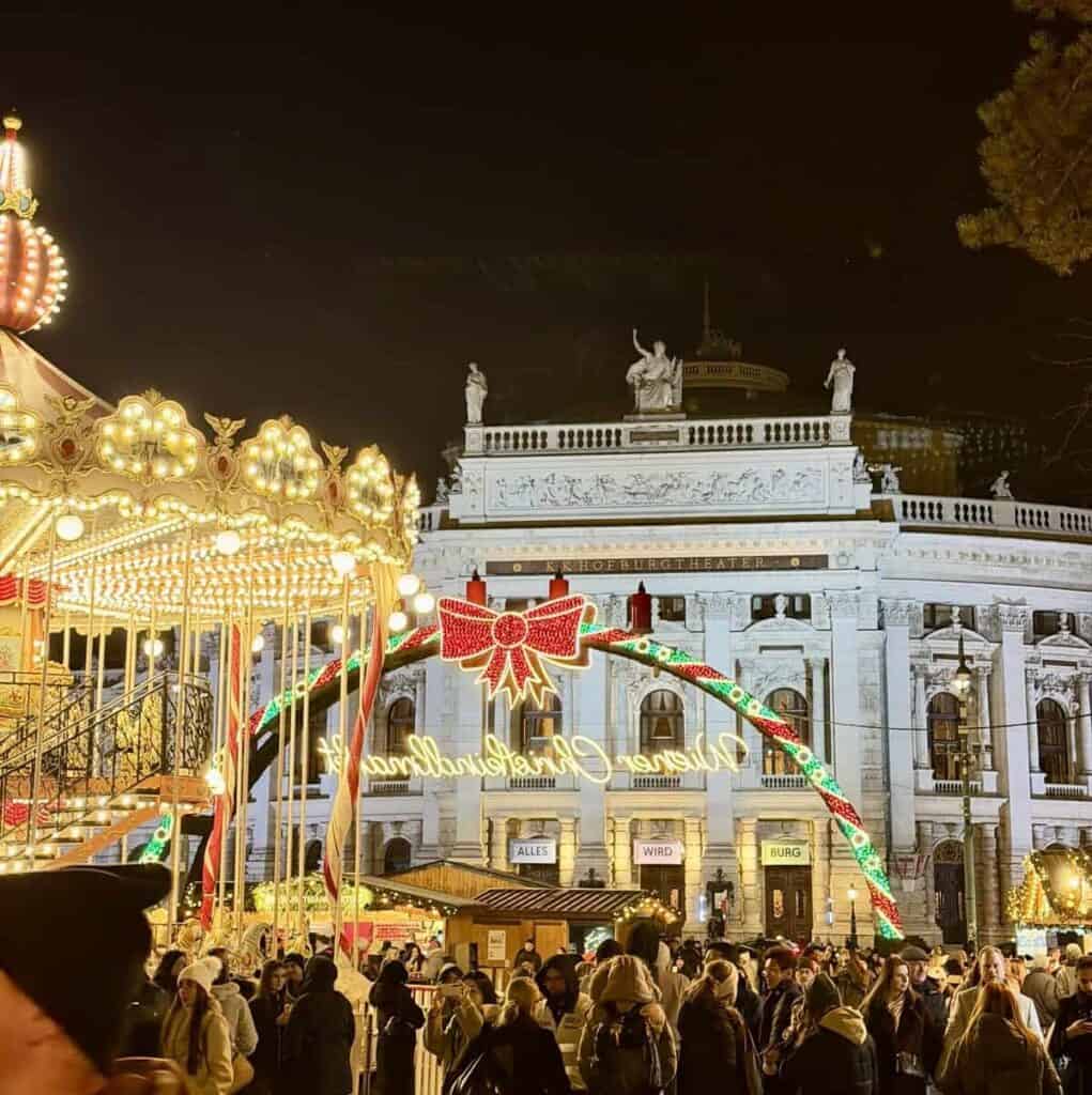 A crowded Vienna Christmas market glows at night beneath a colorful illuminated arch decorated with a red bow. A brightly lit carousel stands beside the historic Burgtheater as visitors explore the festive stalls.