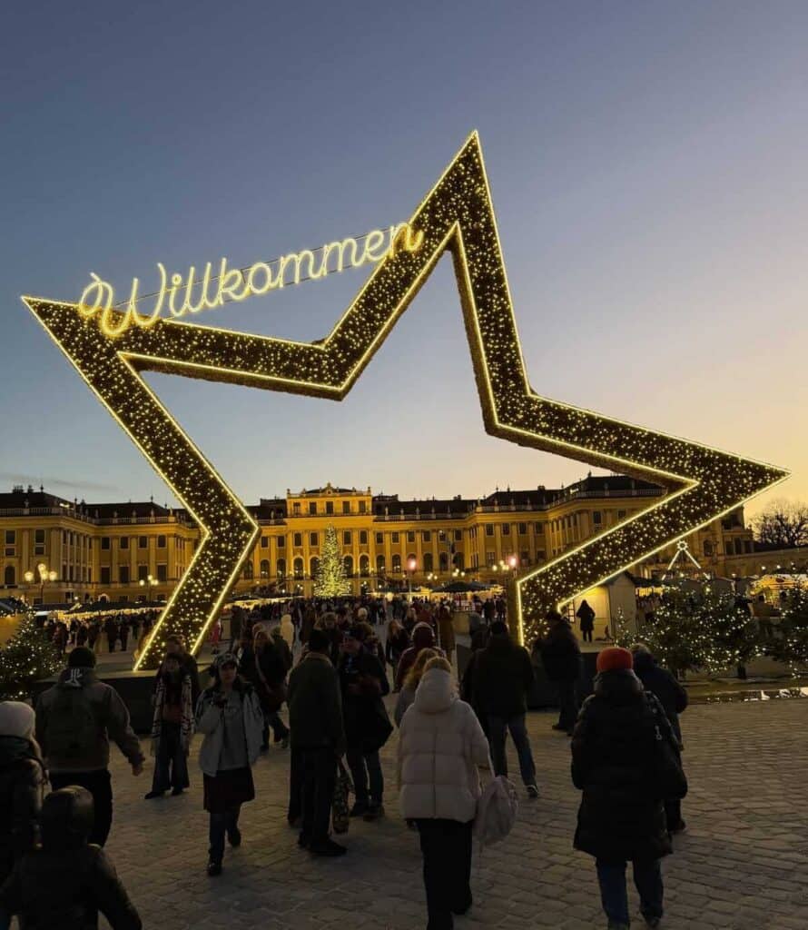 A giant illuminated star with the word "Willkommen" welcomes visitors to the Christmas market at Schönbrunn Palace in Vienna. Crowds walk beneath the golden lights toward the palace and festive market stalls at dusk.