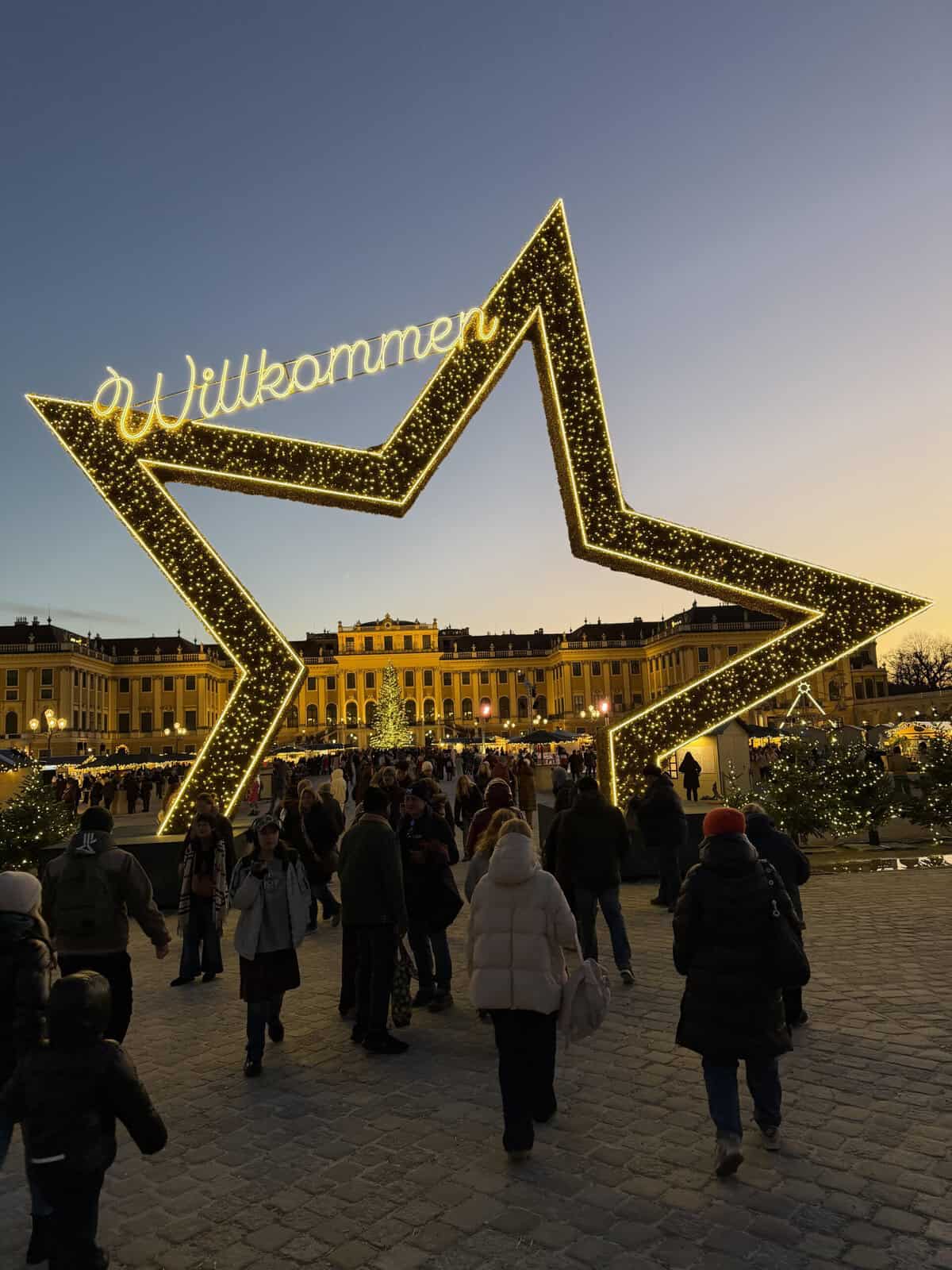 A giant illuminated star with the word "Willkommen" welcomes visitors to the Christmas market at Schönbrunn Palace in Vienna. Crowds walk beneath the golden lights toward the palace and festive market stalls at dusk.